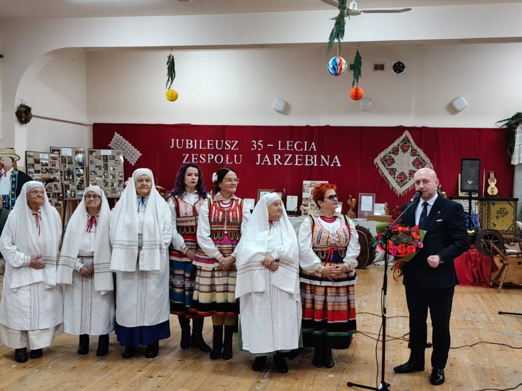 Grupa osób w tradycyjnych strojach ludowych stoi na tle czerwonej ściany z napisem jubileusz 35-lecia zespołu Jarzębina, mężczyzna w garniturze trzyma bukiet kwiatów i mówi do mikrofonu.