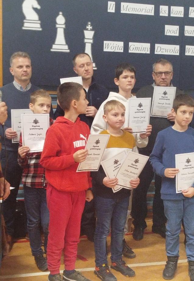 Grupa chłopców i mężczyzn stojących na sali gimnastycznej, każdy trzyma dyplom, w tle plansza z dużymi symbolami figur szachowych i tekstem.