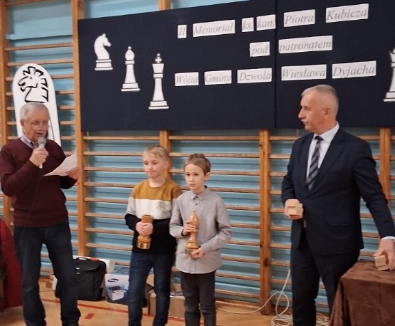 Cztery osoby stojące w sali gimnastycznej, dwie dzieci trzymające nagrody, mężczyzna z mikrofonem i mężczyzna w garniturze z drewnianymi przedmiotami, plansza z szachowymi figurami i tekstem w tle.