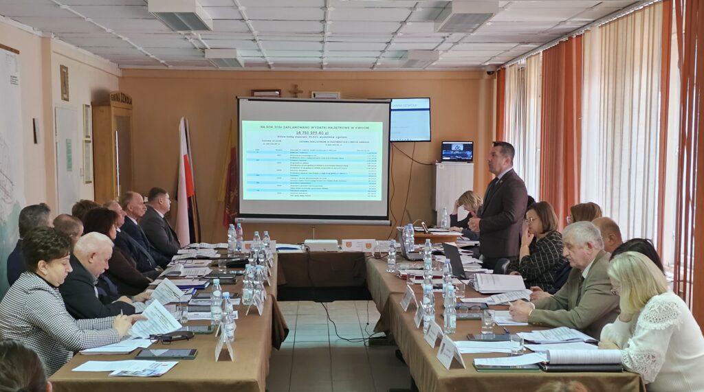 Sala konferencyjna z dwoma rzędami stołów, na których leżą dokumenty i butelki wody, ludzie siedzą i słuchają mężczyzny w garniturze przemawiającego przy ekranie z prezentacją.