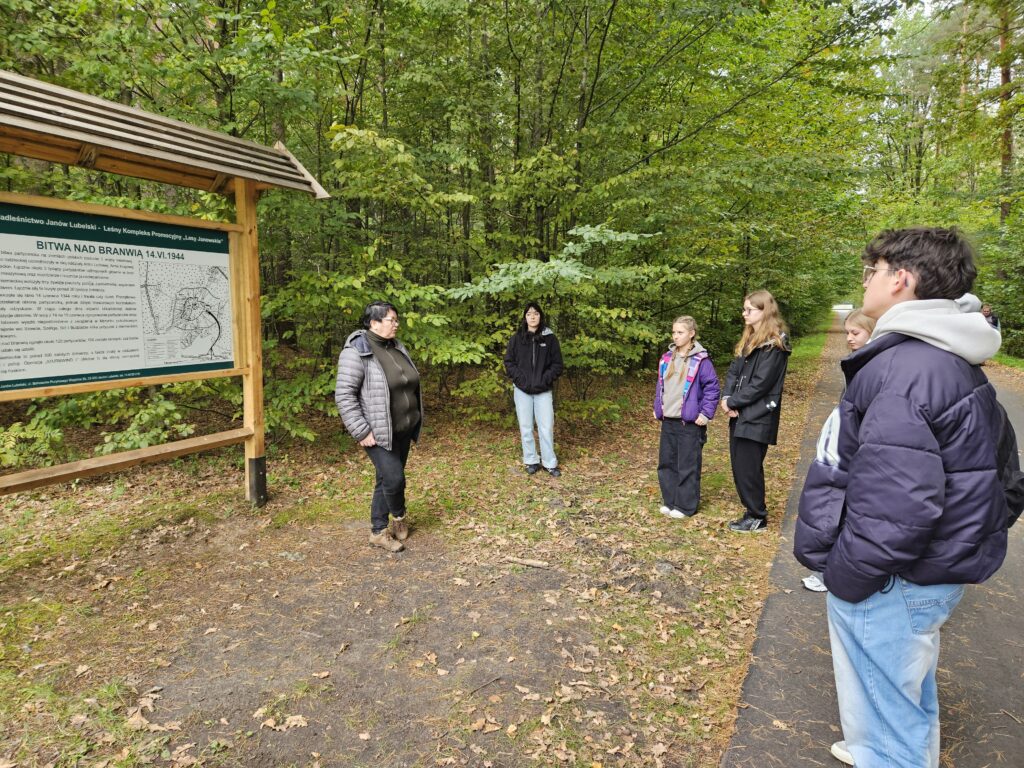 Grupa młodych osób w kurtkach stoi na leśnej ścieżce obok tablicy informacyjnej z mapą i tekstem, otoczeni zielonymi drzewami.