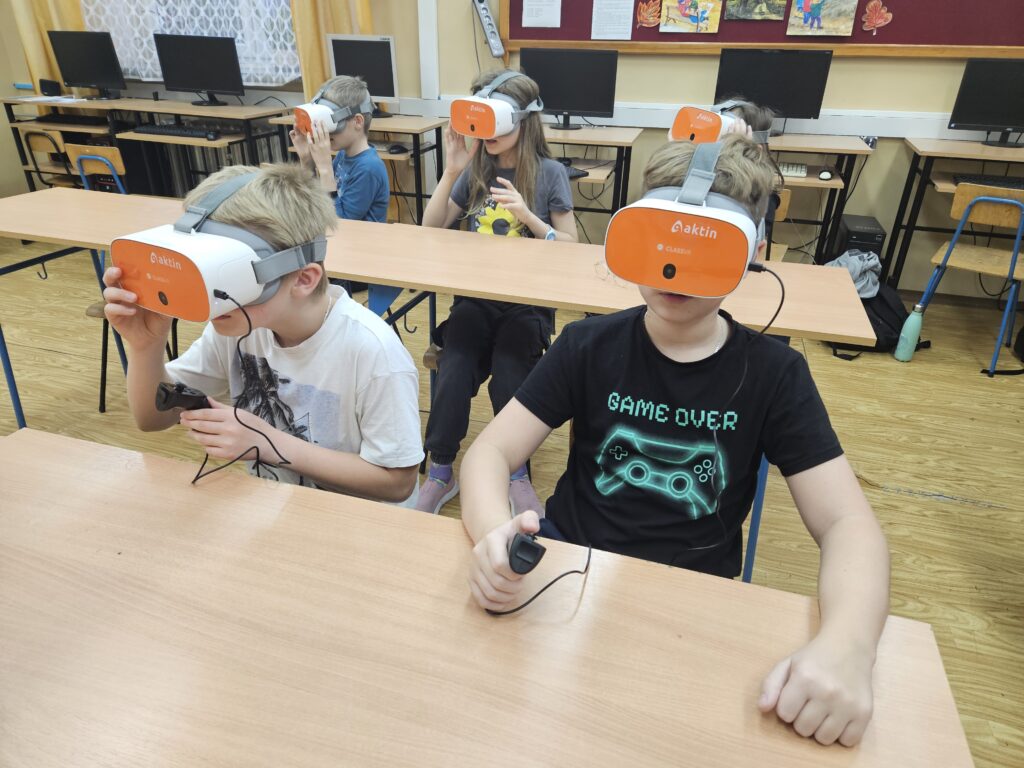 Cztery dzieci siedzące przy drewnianych stołach w klasie, każde w słuchawkach VR z pomarańczowo-białymi goglami, kontrolery w rękach, komputery i tablica w tle.
