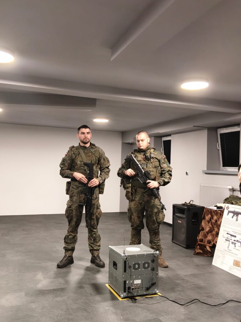 Dwóch żołnierzy w mundurach kamuflażowych stoi obok metalowego urządzenia na szarym podłogowym tle w jasnym pomieszczeniu.