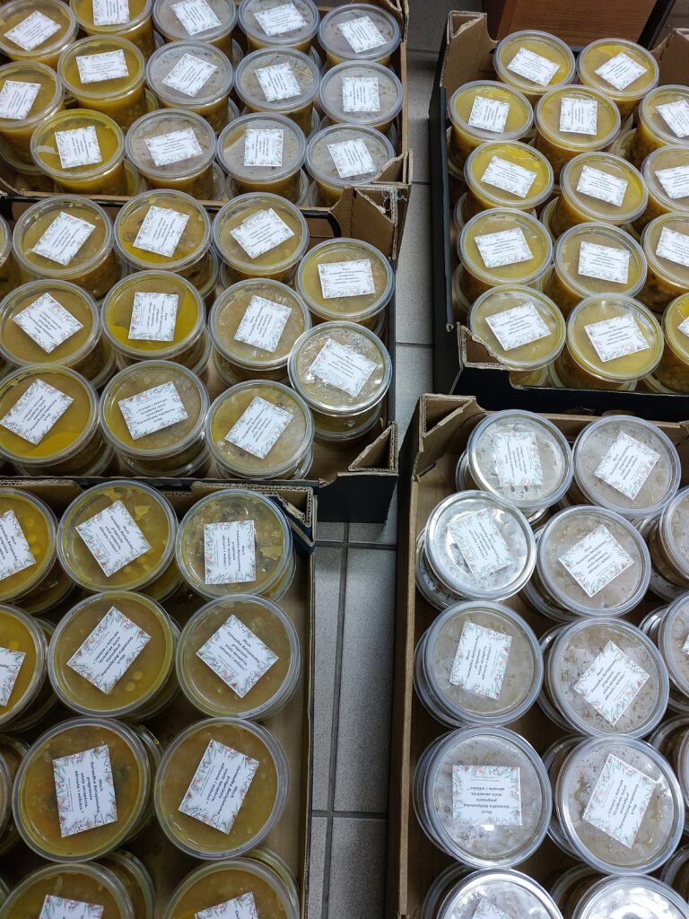 Plastikowe pojemniki z różnokolorową zawartością, ułożone w kartonowych pudełkach na jasnoszarej podłodze. Każdy pojemnik ma naklejkę z tekstem.