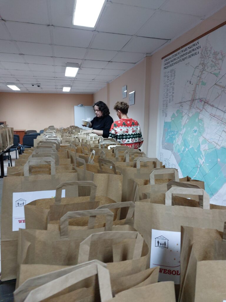 Wiele brązowych papierowych toreb z uchwytami na stołach, dwie osoby przy torebkach, ściana z dużą mapą, jasne oświetlenie sufitowe, jasnoróżowe ściany.