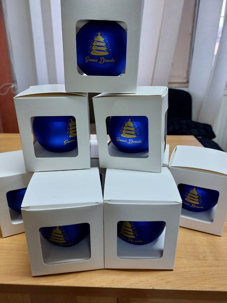 Siedem niebieskich bombek świątecznych z złotym wzorem choinki i napisem "Gmina Dzwola" w białych pudełkach z przezroczystym okienkiem na drewnianym stole.