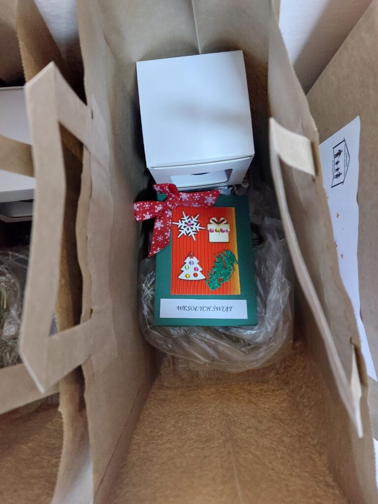 Brązowa torba papierowa z białym pudełkiem i zieloną kartką świąteczną z czerwoną kokardą i ozdobami, napis „WESOŁYCH ŚWIĄT”.