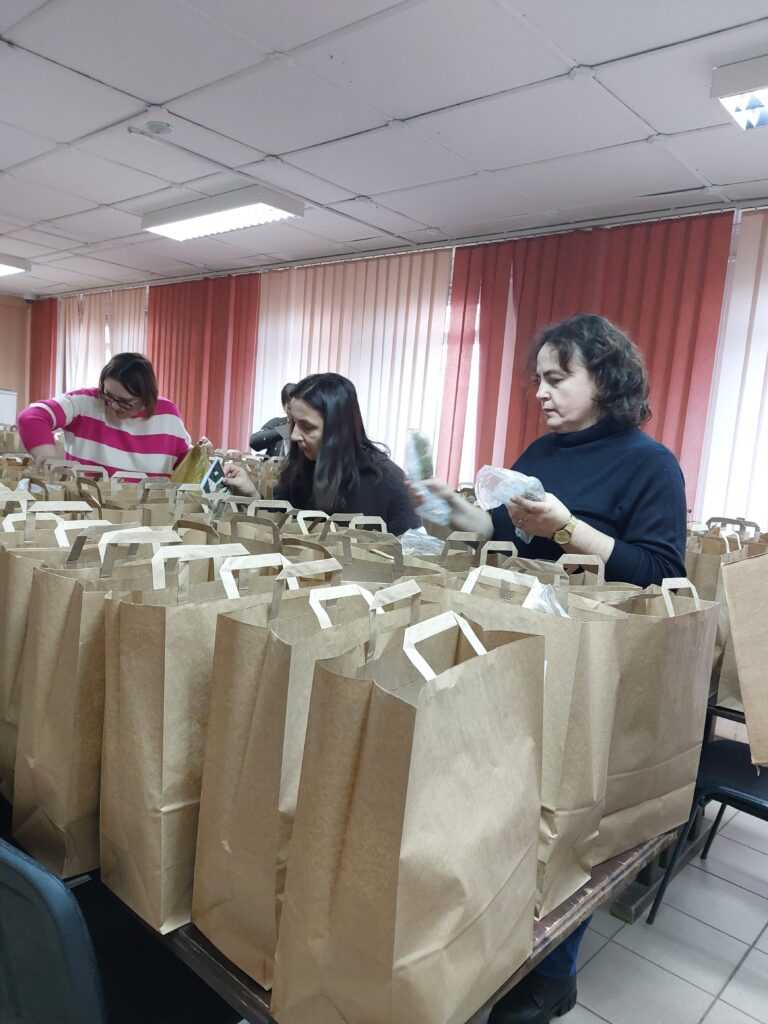 Kobiety pakujące produkty do brązowych papierowych toreb na stole w pomieszczeniu z czerwono-beżowymi zasłonami i białym sufitem.