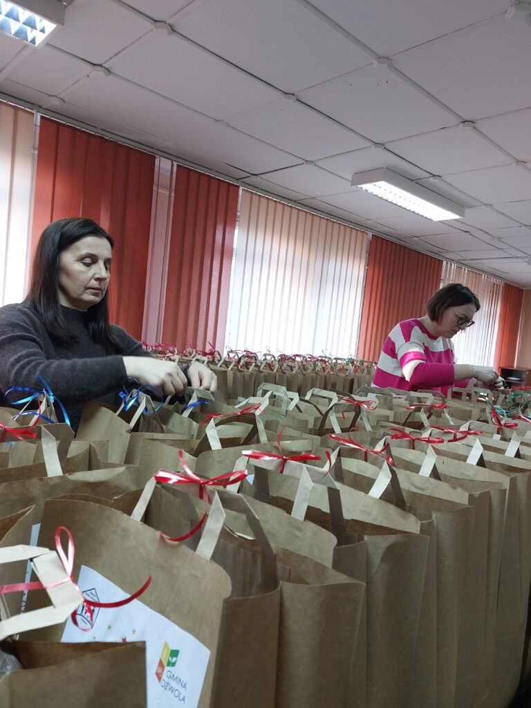 Dwie kobiety przy stole obwiązywające brązowe torby papierowe czerwoną i niebieską wstążką, za nimi czerwone i białe pionowe żaluzje.