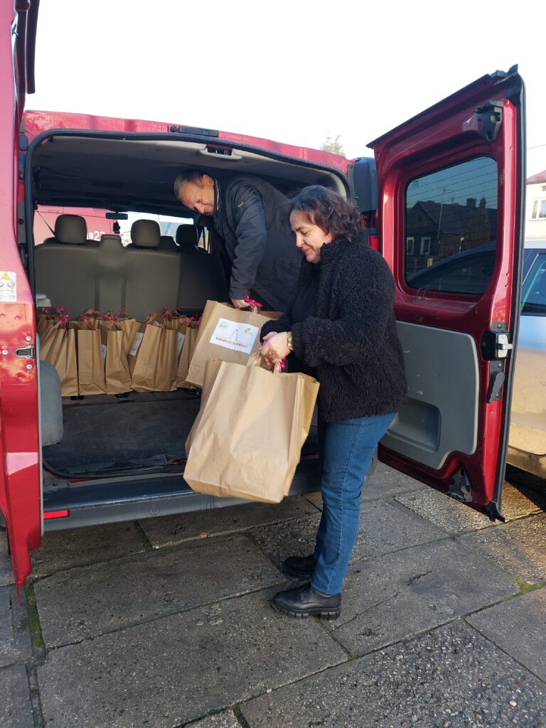 Czerwony pojazd z otwartymi tylnymi drzwiami, mężczyzna w czarnej kurtce wkładający papierową torbę do środka, kobieta w czarnym swetrze i niebieskich spodniach trzymająca dużą papierową torbę.