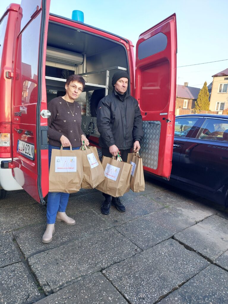 Kobieta i mężczyzna trzymają torby papierowe z napisami „Wesołych Świąt” stojąc przy otwartych drzwiach czerwonego pojazdu na tle budynków i samochodu.