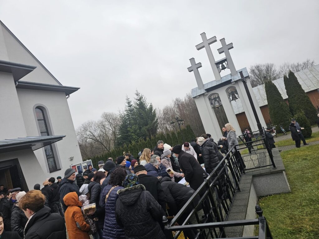 Grupa ludzi w zimowych ubraniach zgromadzona przed budynkiem z szarymi ścianami, obok trzech dużych białych krzyży z dzwonami, zielona trawa i drzewa w tle.