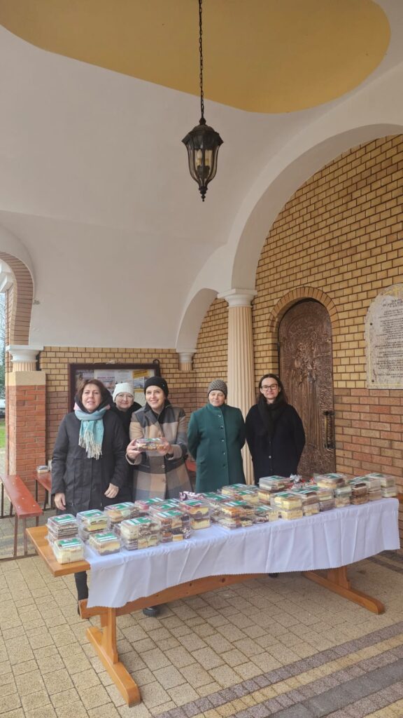 Pięć kobiet w kurtkach i czapkach stoi za stołem z białym obrusem, na którym ułożone są plastikowe pudełka z ciastami, podcienie i żółto-brązowa ceglana ściana.