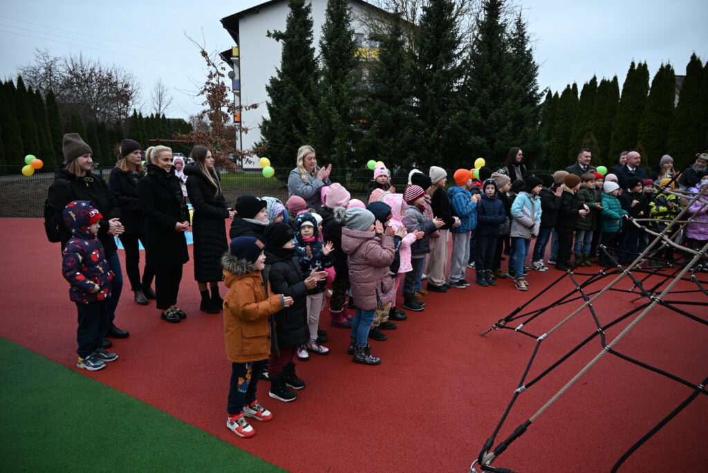 Grupa dzieci w kolorowych kurtkach i czapkach stojących na czerwonym placu zabaw, w tle dorosłe osoby i zieleń drzew iglastych.
