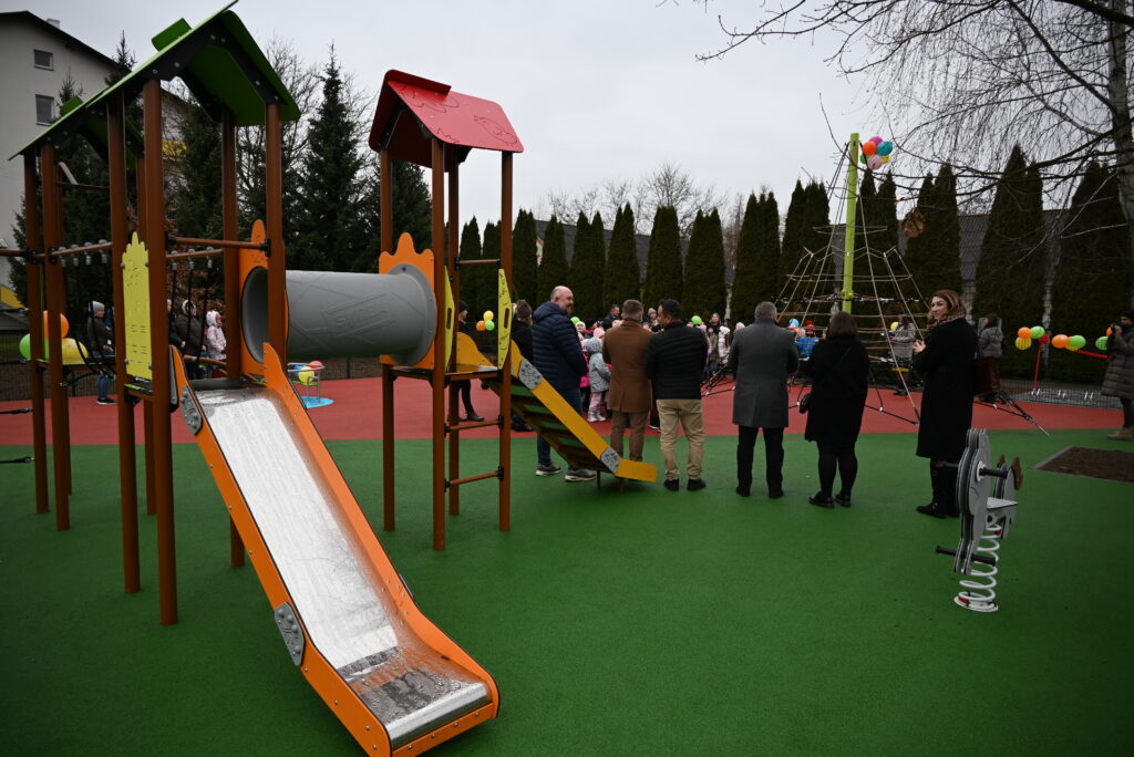 Plac zabaw z zieloną i czerwoną nawierzchnią, zjeżdżalnia z pomarańczowymi bokami, drabinka, domek z czerwonym i zielonym dachem, ludzie stojący w grupie, drzewo bez liści.
