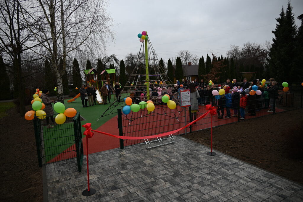 Plac zabaw z linową konstrukcją i zjeżdżalnią, ogrodzony balonami i czerwoną wstęgą, wokół zgromadzeni dorośli i dzieci w kolorowych ubraniach, pochmurne niebo.