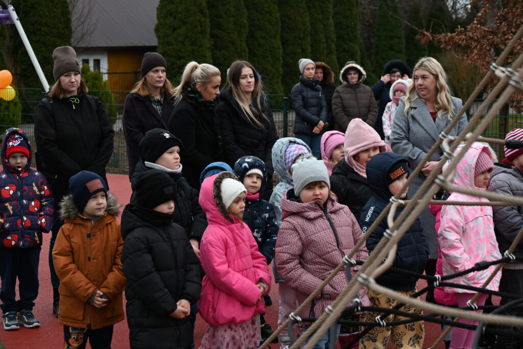 Grupa dzieci i dorosłych ubranych w zimowe kurtki i czapki stoi na placu zabaw z czerwonym podłożem, w tle zielone drzewa i ogrodzenie.