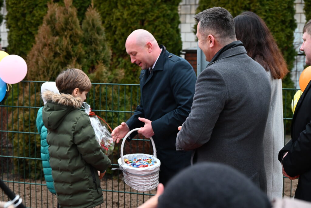 Mężczyzna w granatowym płaszczu podaje koszyk z cukierkami chłopcu w zielonej kurtce, w tle trzy osoby i zielony metalowy płot, balony po lewej stronie.
