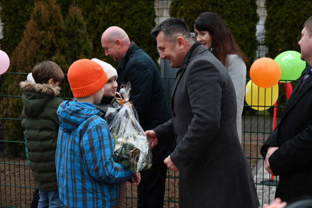 Dzieci w kurtkach i czapkach przekazują mężczyźnie w szarym płaszczu prezent w przezroczystym opakowaniu, w tle kolorowe balony i osoby na zewnątrz przy ogrodzeniu.