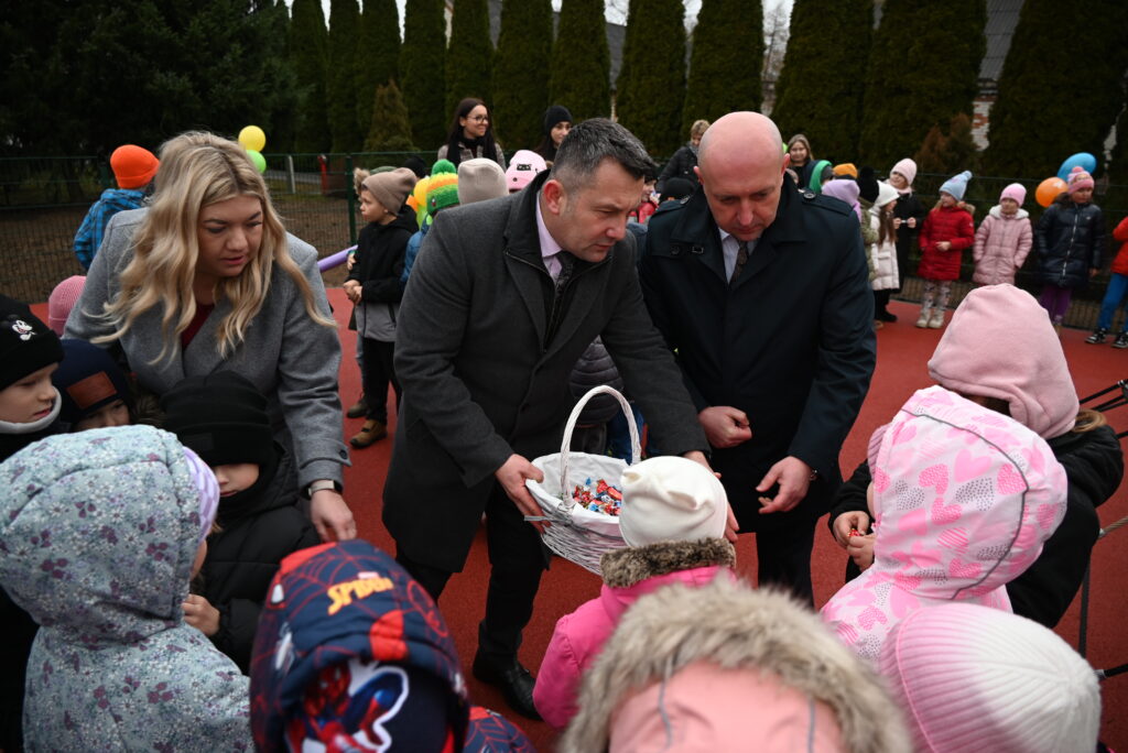 Dwóch mężczyzn w płaszczach rozdaje cukierki z wiklinowego kosza grupie dzieci ubranych w kolorowe kurtki i czapki na tle czerwonej nawierzchni i zielonych drzew.