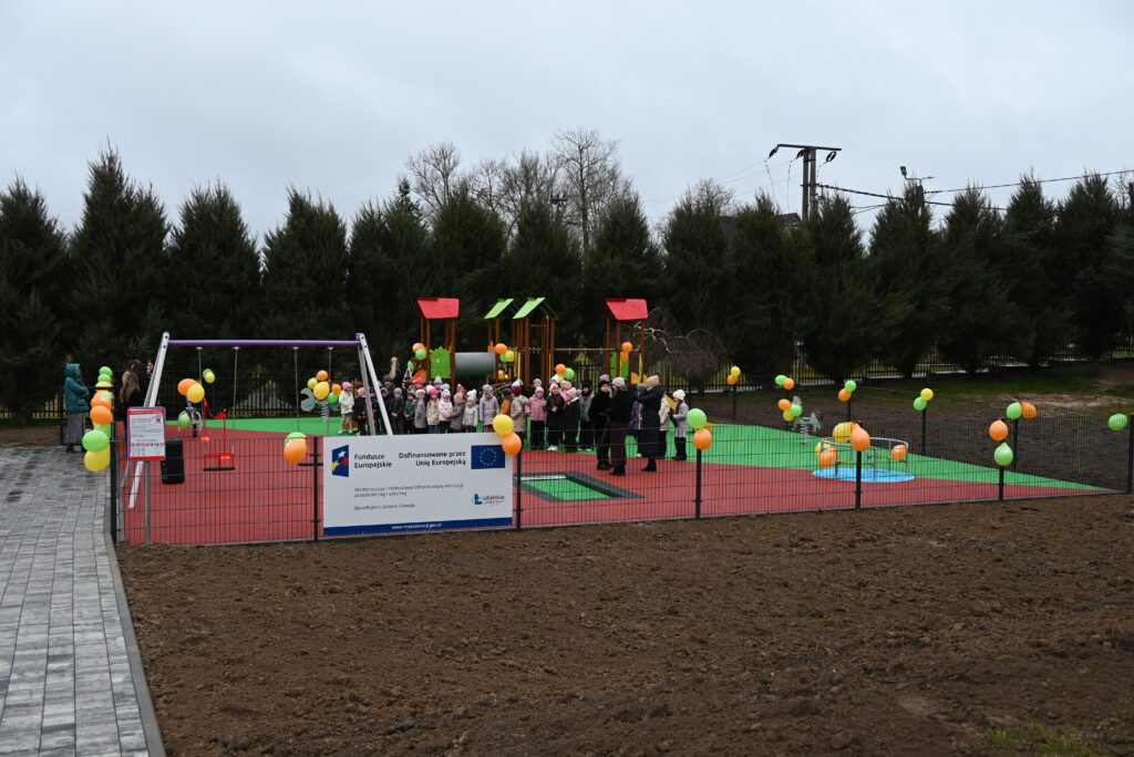 Plac zabaw z huśtawkami i zjeżdżalniami, ogrodzony metalowym płotem, z kolorowymi balonami, grupą dzieci i dorosłych, w tle wysokie zielone drzewa, pochmurne niebo.