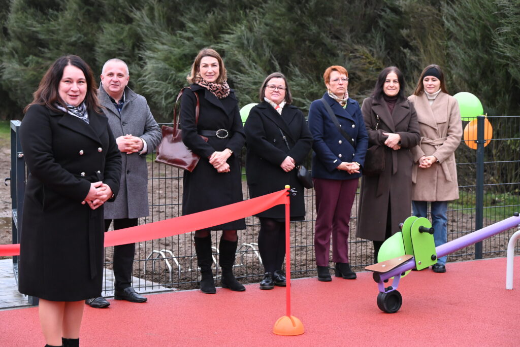 Siedem osób w płaszczach stoi na czerwonej nawierzchni placu zabaw, za nimi metalowe ogrodzenie i zielone drzewa, z przodu huśtawka ważka i czerwona wstęga.
