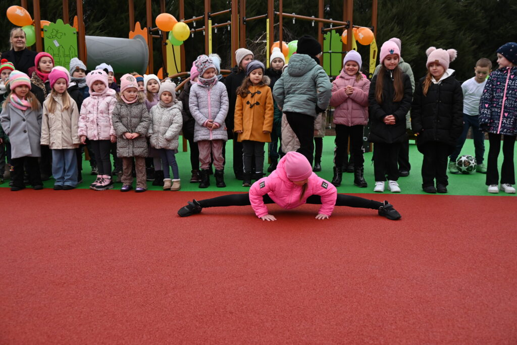 Dziecko w różowej kurtce i czapce wykonuje szpagat na czerwonym podłożu, w tle stoi grupa dzieci w kolorowych zimowych ubraniach na placu zabaw z balonami.