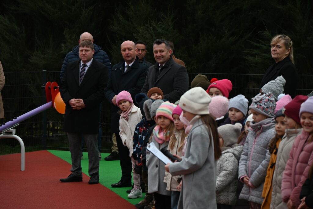 Grupa dzieci w kolorowych czapkach i kurtkach stoi w rzędzie obok trzech mężczyzn w garniturach i jednej kobiety na tle zielonych drzew i ogrodzenia.