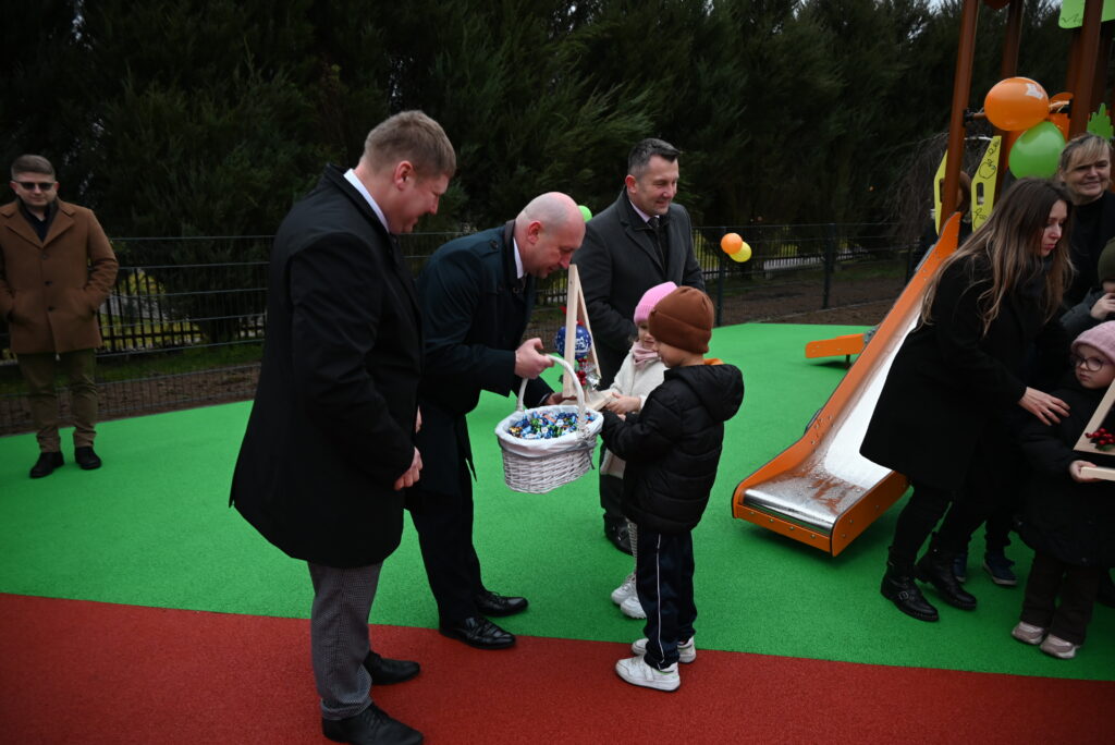 Mężczyźni w ciemnych płaszczach wręczają dzieciom kosz z cukierkami na placu zabaw z zieloną i czerwoną nawierzchnią oraz metalową zjeżdżalnią.