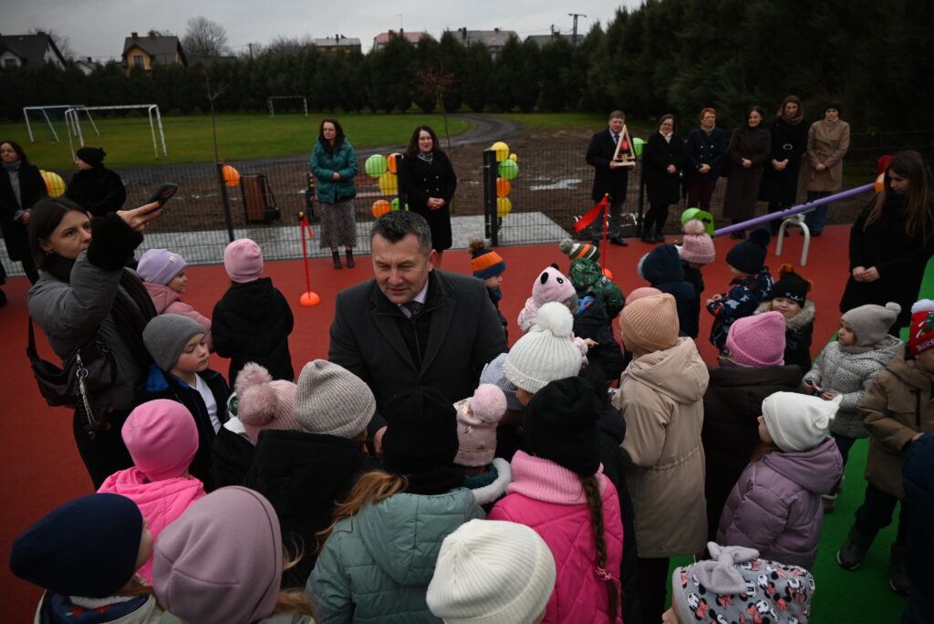 Duża grupa dzieci w kolorowych kurtkach i czapkach oraz dorośli na tle boiska z czerwoną i zieloną nawierzchnią, w tle drzewa i budynki mieszkalne.