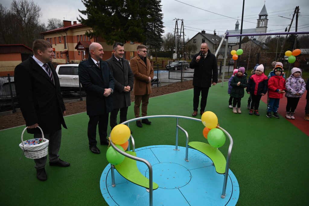 Mężczyźni w płaszczach obok karuzeli z balonami, dzieci ustawione w rzędzie na zielonej nawierzchni placu zabaw, w tle budynki i szare niebo.