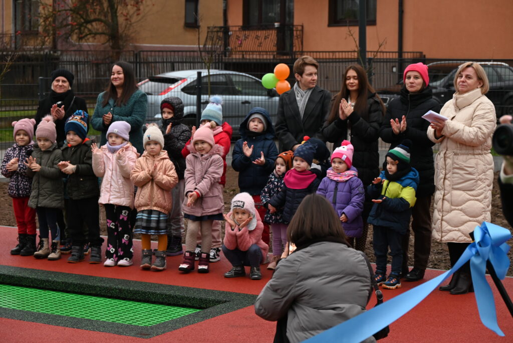 Grupa dzieci i dorosłych ubranych w kolorowe kurtki i czapki stoi na zewnętrznym placu zabaw z czerwonym i zielonym podłożem, obok błękitnej wstążki.