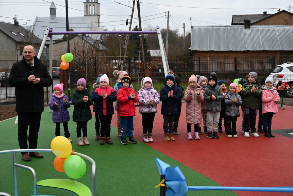 Grupa dzieci ubranych w kolorowe kurtki i czapki stoi na placu zabaw z zieloną i czerwoną nawierzchnią, obok stoi mężczyzna w czarnym płaszczu z mikrofonem.
