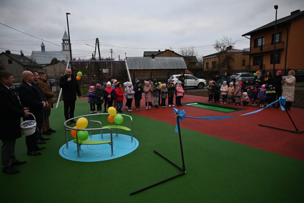 Dzieci i dorośli stojący na zielono-czerwonym placu zabaw, balony przy huśtawce i karuzeli, w tle domy i kościół pod pochmurnym niebem.