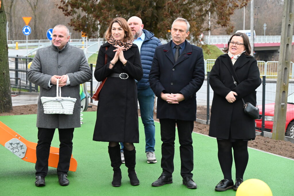 Cztery osoby w ciemnych płaszczach stoją na zielonej nawierzchni na zewnątrz, mężczyzna z lewej trzyma białe wiklinowe wiadro, w tle drzewa i ogrodzenie.