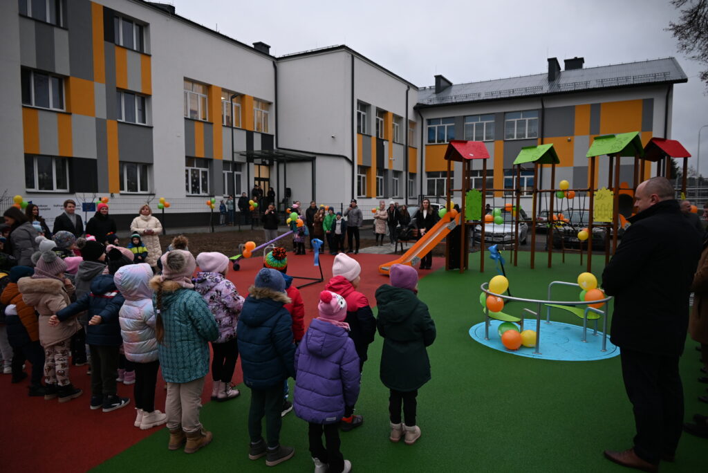 Dzieci w kolorowych kurtkach stojące na placu zabaw z zieloną i czerwoną nawierzchnią, z huśtawką, zjeżdżalnią i karuzelą, w tle budynek z szaro-pomarańczowymi panelami.