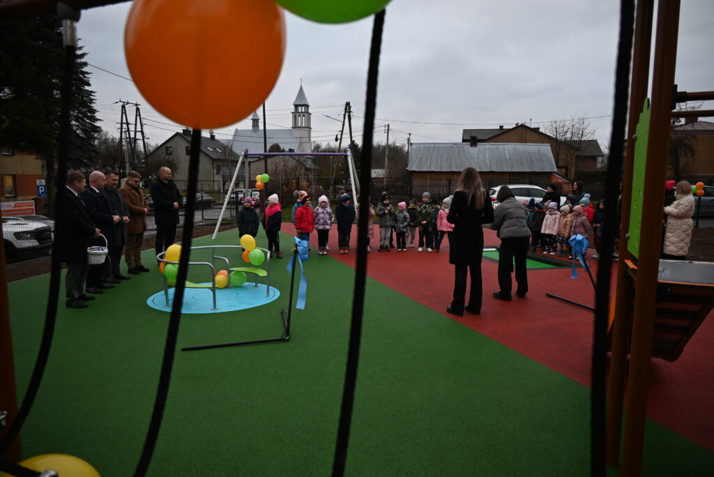 Dzieci i dorośli stoją na kolorowym placu zabaw z zieloną i czerwoną nawierzchnią, wokół balony w kolorach pomarańczowym, zielonym i żółtym, w tle domy i kościół.