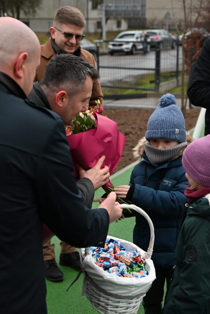 Dzieci w zimowych kurtkach i czapkach odbierają bukiet kwiatów i kosz pełen kolorowych słodyczy od dorosłych mężczyzn na zielonym placu.