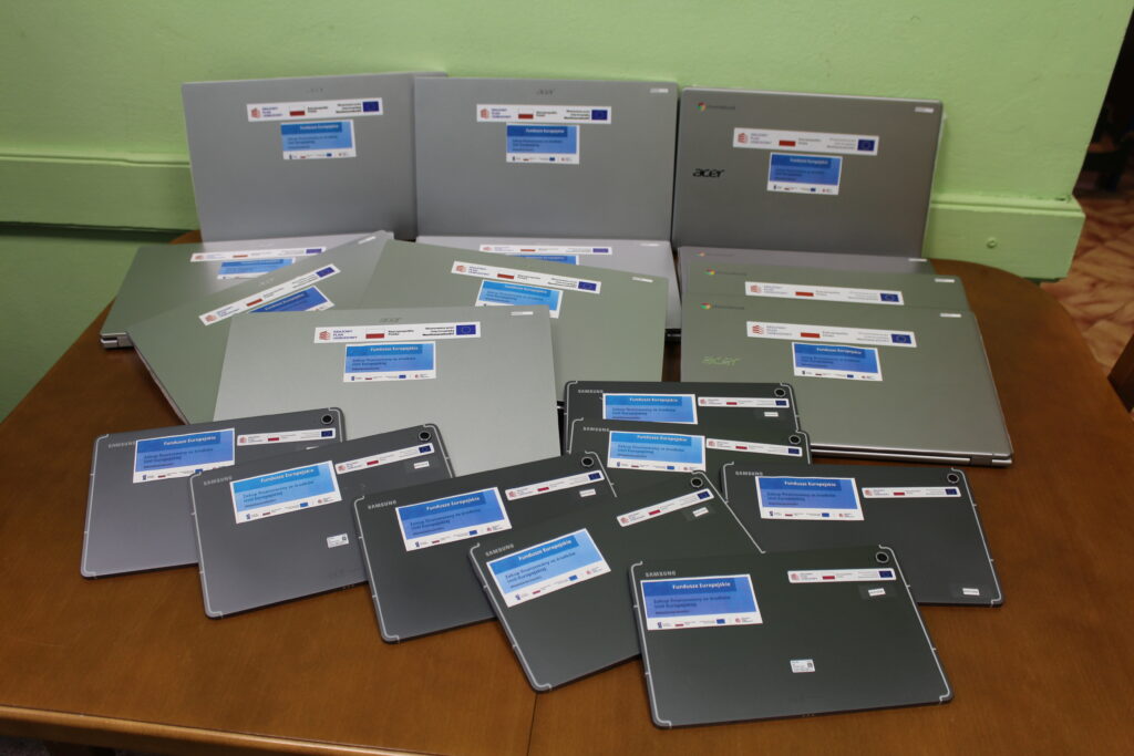 Laptop i tablety w różnych rozmiarach, szare i srebrne, ułożone na drewnianym stole, z naklejkami informacyjnymi o funduszach europejskich.