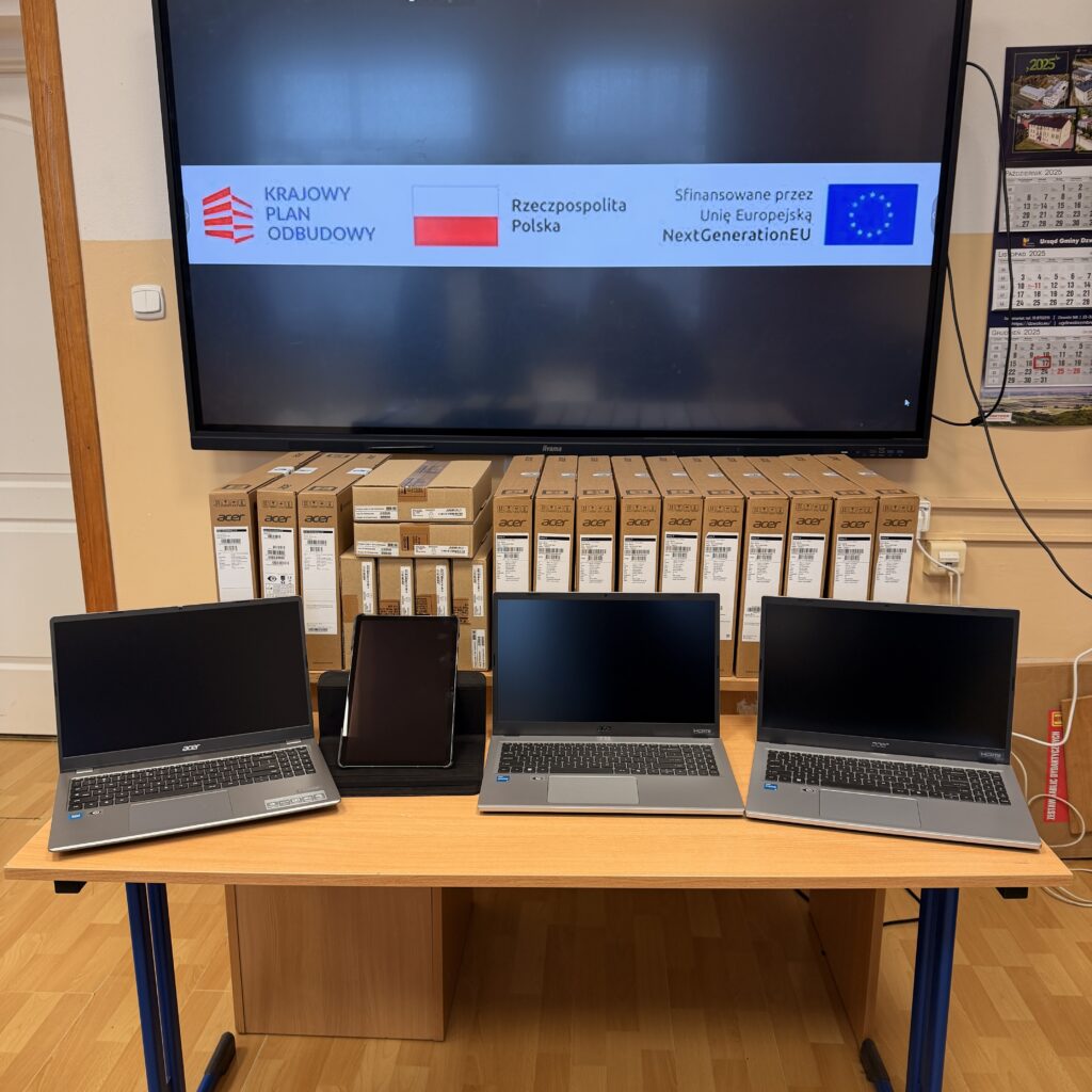 Trzy srebrne laptopy Acer i czarna tabletka na stojaku na drewnianym stole, za nimi stos kartonowych pudełek, na ekranie napis "Krajowy Plan Odbudowy", flaga Polski i Unii Europejskiej.