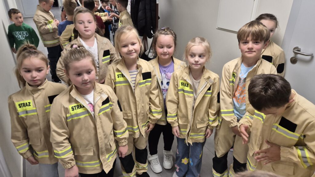 Grupa dzieci w beżowych kurtkach strażackich z żółtymi odblaskowymi paskami stoi w korytarzu o szarych ścianach i podłodze.