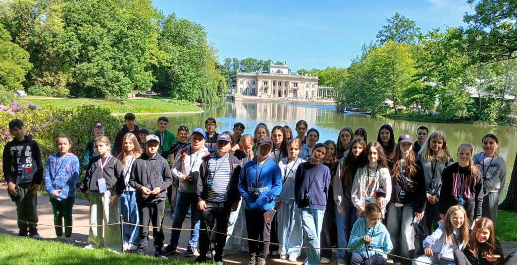 Grupa dzieci na ścieżce nad jeziorem z zielonymi drzewami i białym pałacem w tle, niebieskie niebo.