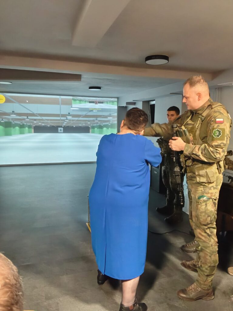 Kobieta w niebieskiej sukience celuje z broni pod okiem żołnierza w mundurze moro, w tle ekran z symulacją strzelnicy, drugi żołnierz obserwuje.