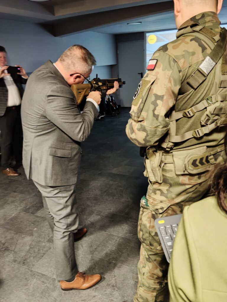 Mężczyzna w szarym garniturze celuje z broni, obok stoi żołnierz w mundurze kamuflażowym z polską flagą na ramieniu, w tle mężczyzna robiący zdjęcie telefonem.