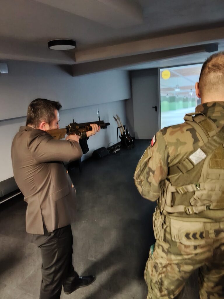 Mężczyzna w garniturze celuje z karabinu, obok stoi żołnierz w kamuflażu, w tle białe ściany i ciemna podłoga, na ścianie widoczny ekran projekcyjny.