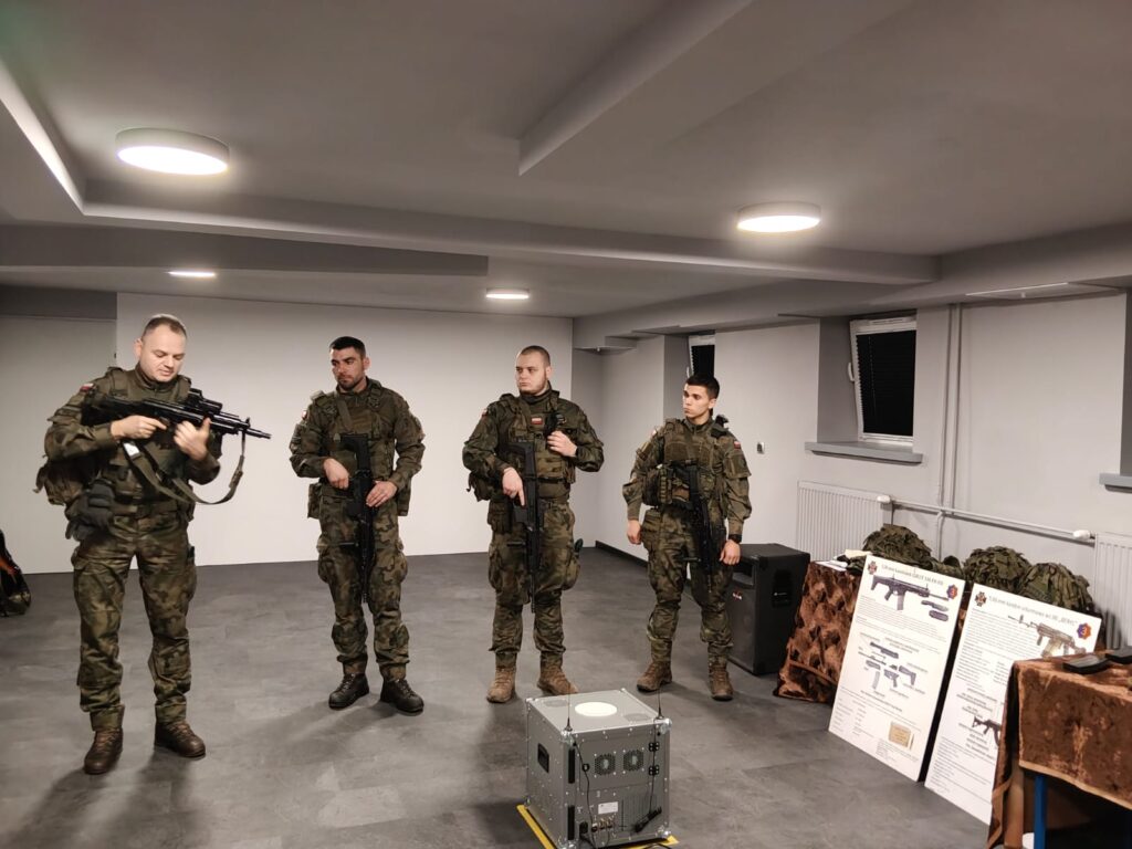 Czterech żołnierzy w zielonym umundurowaniu i brązowych butach stoi w jasnym pomieszczeniu z szarym podłogą, obok stołów z plakatami i sprzętem wojskowym.