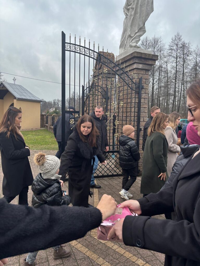 Grupa ludzi w ciemnych płaszczach na tle ozdobnej czarnej bramy i pomnika, w tle szare niebo i drzewa bez liści.
