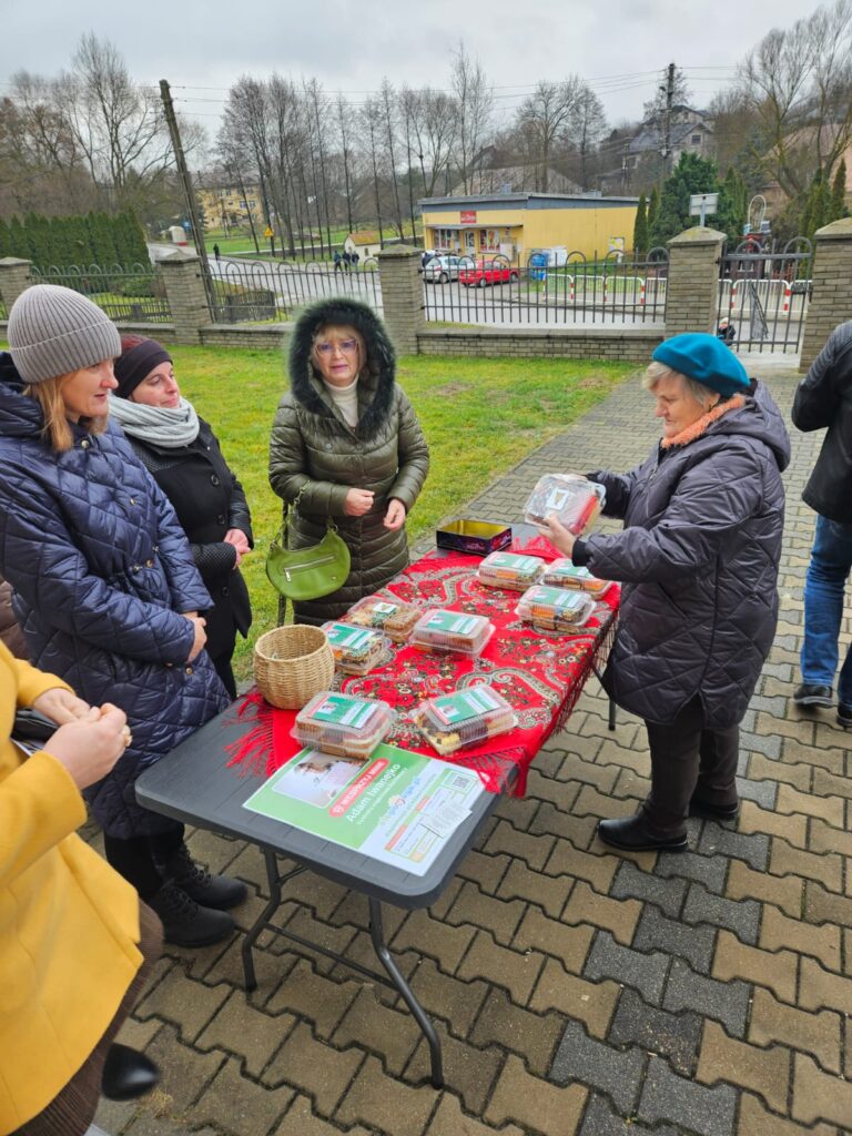 Grupa kobiet w kurtkach zimowych stoi przy stole z czerwonym obrusem, na którym leżą plastikowe pojemniki z ciastem i koszyk, w tle płot i budynki.