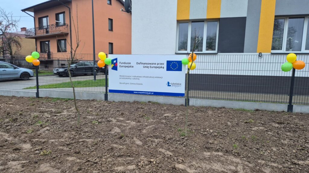 Teren z gołą ziemią i dwoma cienkimi drzewkami przy ogrodzeniu z balonami w kolorach zielonym, żółtym i pomarańczowym, na tle budynków i tablica informacyjna.