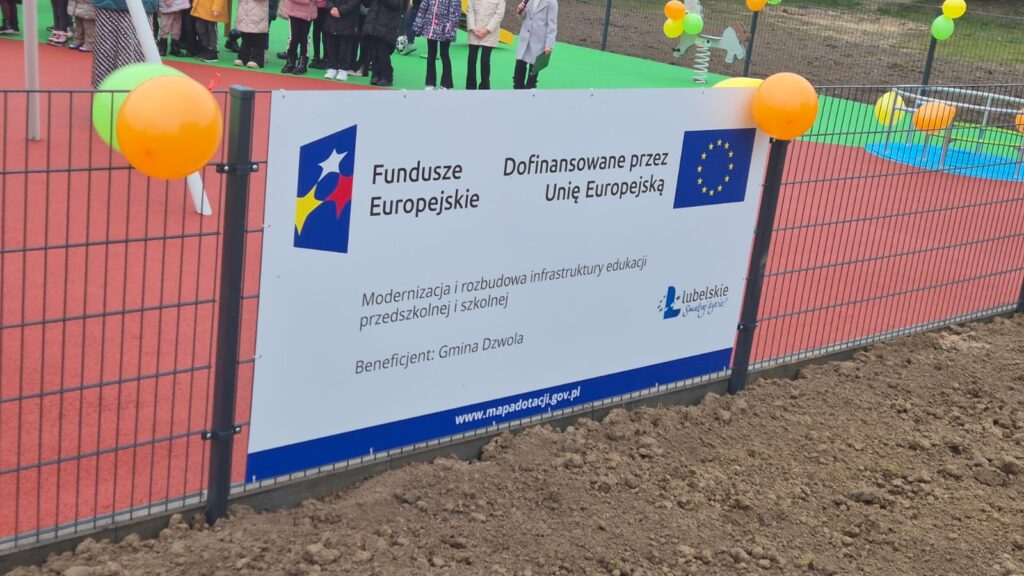 Tablica informacyjna o dofinansowaniu z Unii Europejskiej na ogrodzeniu wokół placu zabaw z czerwoną i zieloną nawierzchnią, przy tablicy pomarańczowe i zielone balony.